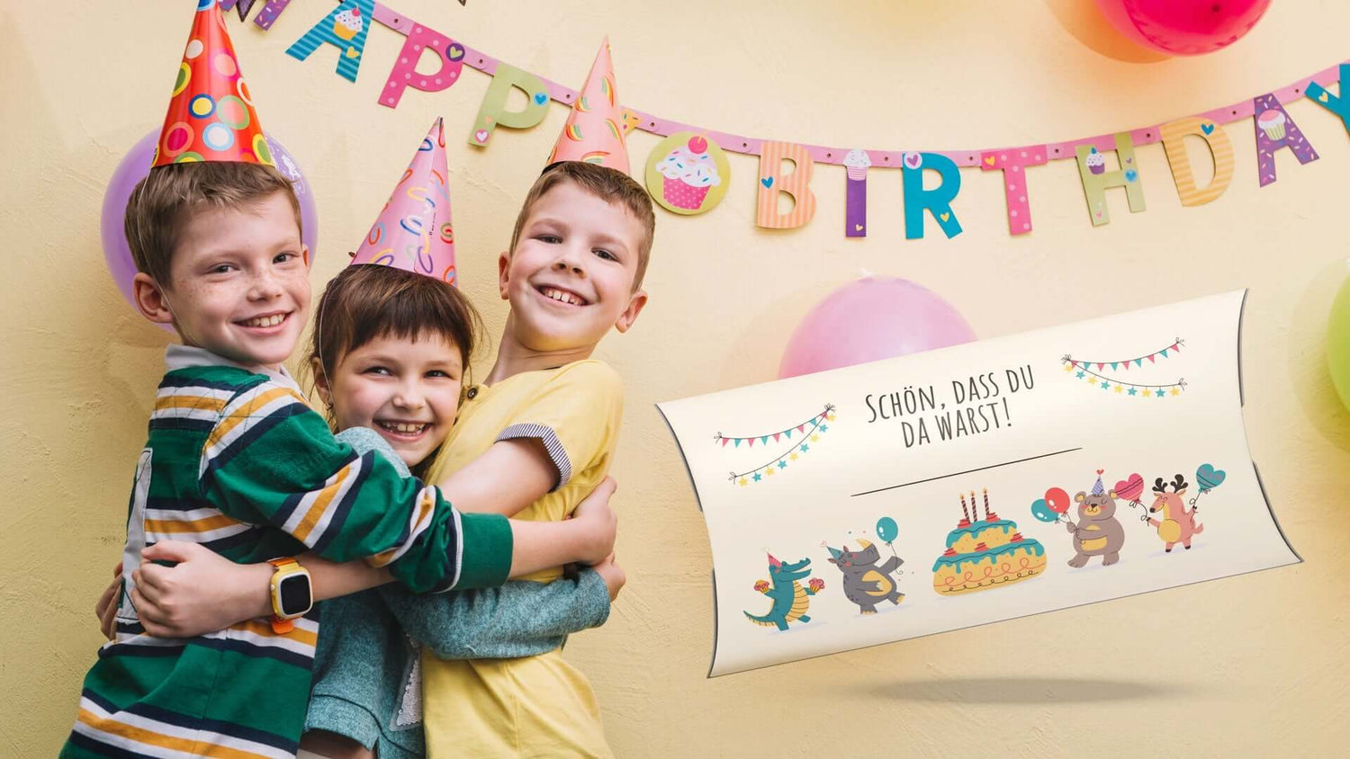 Drei fröhliche Kinder mit Geburtstagshüten umarmen sich vor einer Geburtstagsbanner-Deko.